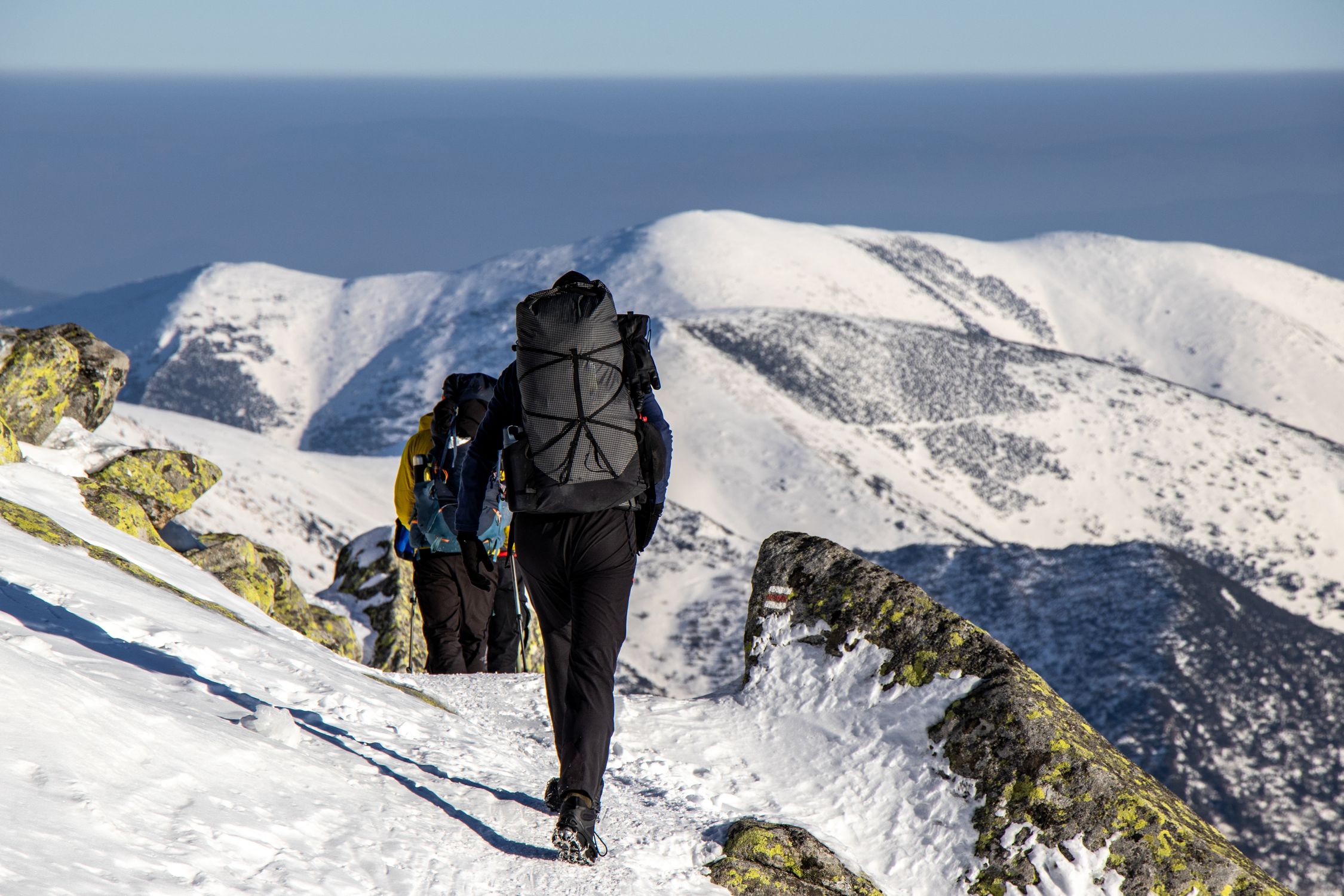 Hikers with backpacks trek through snow-covered mountains on a sunny winter day